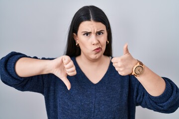 Young brunette woman standing over isolated background doing thumbs up and down, disagreement and agreement expression. crazy conflict