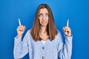 Young woman standing over blue background pointing up looking sad and upset, indicating direction with fingers, unhappy and depressed.