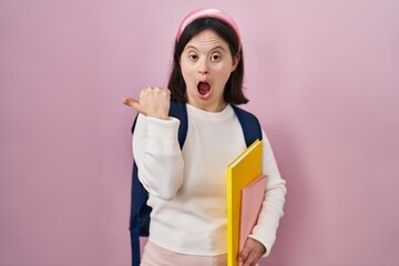 Woman with down syndrome wearing student backpack and holding books surprised pointing with hand...
