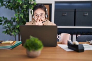 Woman with down syndrome working at the office wearing headset covering mouth with hand, shocked...