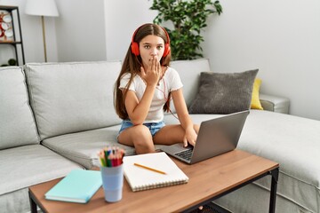 Young brunette teenager doing homework with laptop at home covering mouth with hand, shocked and...