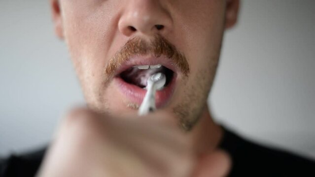 Caucasian man brushing teeth with open mouth in front of white wall. Static close up shot with blurry background