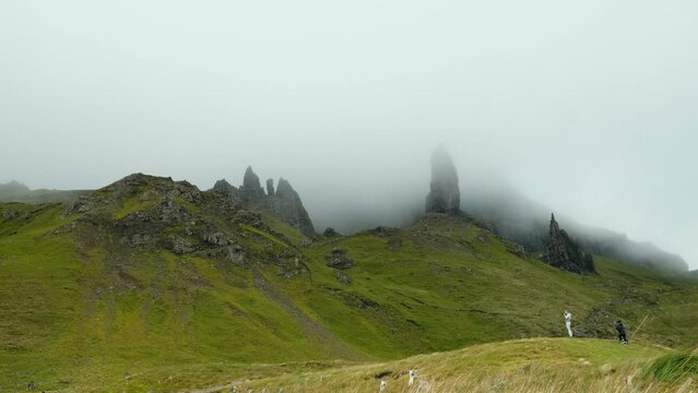 A group of rock formations beneath the mountains, immersed in heavy fog. some distant unrecognized people are visible, taking pictures and enjoying the view. 4K video clip, old man of storr, Scotland.