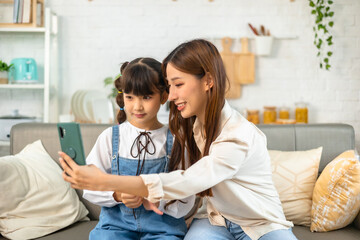 Obraz premium Young asian mother and her daughter are making selfie using a phone, hugging and smiling while sitting on the sofa at home.