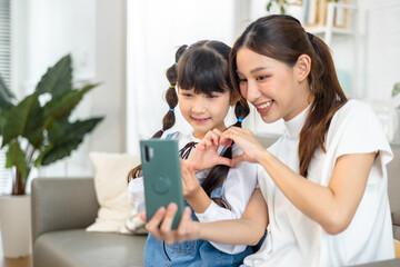 Young asian mother and her daughter are making selfie using a phone, hugging and smiling while sitting on the sofa at home.