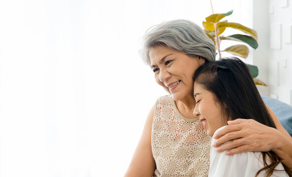 Portrait of happy Asian senior, mother white hair embracing her beautiful daughter with love, care, comfort and smile while sit on sofa on white background in living room at home with copy space.