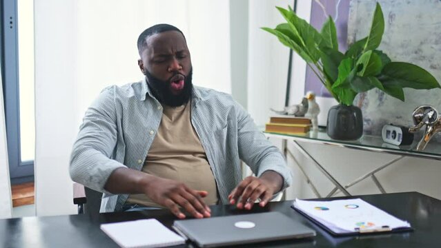 End of the working day. Overjoyed happy cheerful african american young man sitting in the office at the table, closes the laptop, finishes work, dancing with joy, smiles, gesticulates with hands