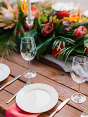 Decor and serving of a wedding banquet on a wooden table. White plates on a red napkin, cutlery, and wine glasses. Tropical flowers in the background.