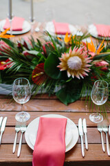 Decor and serving of a wedding banquet in a tropical style. Exotic flowers, protea, strelitzia, leaves, white candles in high candlesticks, wine glasses, red napkins on white plates, cutlery on a wood