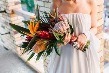 The bride in a dress with bare shoulders holds an exotic wedding bouquet of tropical flowers. Bouquet of protea, strelitzia, red ginger, and tropical leaves. In the background is a brick wall.