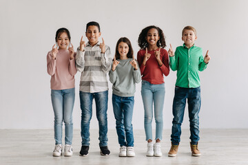 Cheerful Multicultural Preteen Children Pointing Fingers Upward Standing Together Indoors - Powered by Adobe