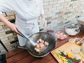 A chef in a white suit fries salmon in a wok. In the background is a wooden board with lemon slices, rosemary, ginger and honey on it.