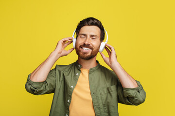 Calm man listening to music with closed eyes, wearing wireless headphones over yellow background