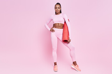 Fototapeta na wymiar Beautiful African woman in sports clothing carrying exercise mat against pink background