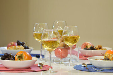 White dining table with white plates full of food and fruit, red grapes, persimmons, nuts and tomatoes, blue and pink placemats and crystal glasses full of white wine