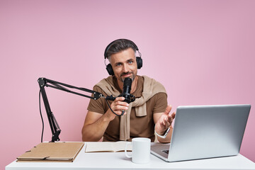 Handsome man in headphones using microphone while recording podcast against pink background