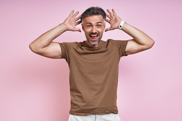 Fototapeta na wymiar Surprised mature man looking at camera and gesturing while standing against pink background