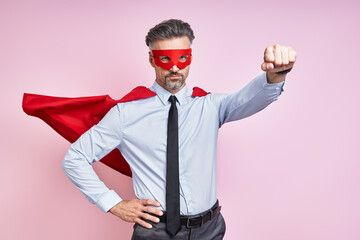 Confident mature man wearing superhero cape and keeping arm outstretched against pink background