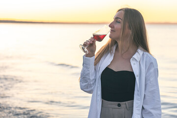 Beautiful woman with a glass of wine against the backdrop of the sea at sunset.