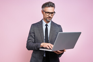 Confident mature man in formalwear using laptop while standing against pink background