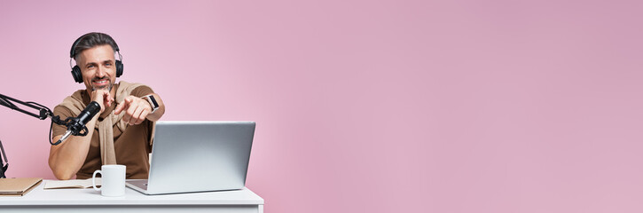 Confident mature man in headphones pointing camera while recording podcast against pink background