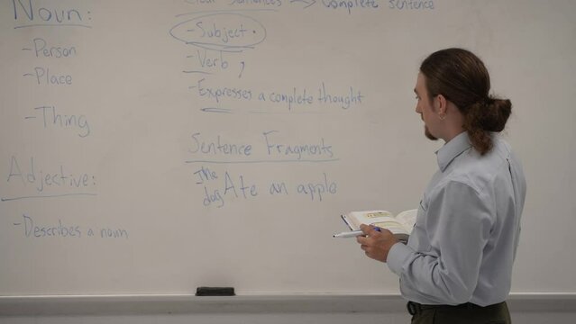 Male school teacher holding book teaching school classroom at a whiteboard, answering questions.