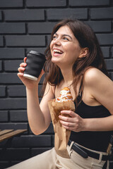 A young woman with a croissant and a cup of coffee against a black brick wall.