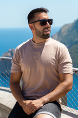 Young man sitting in the foreground with beard and sunglasses with the Mediterranean sea and cliffs in the background.