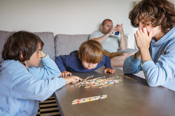 Three brothers playing board game Mikado at home while the father checking his smartphone, family fun, siblings spending time together.