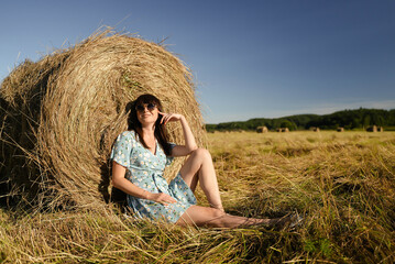 Naklejka na ściany i meble A beautiful girl in a summer dress and sunglasses sits near a haystack and enjoys her vacation.