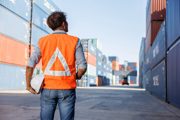 back view engineer or factory worker holding helmet and looking up to containers in warehouse...