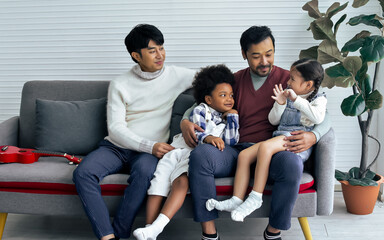 Couple male gay taking care with love to adopted children who are little diverse Caucasian and African girl and boy, sitting on sofa in living room at home. LGBT, kids Concept.
