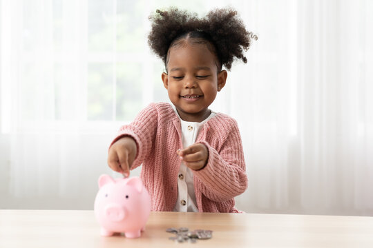 Happy African American small girl kid putting money into the piggybank. Little girl inserting a coin in a piggy bank. Kid investment for future concept