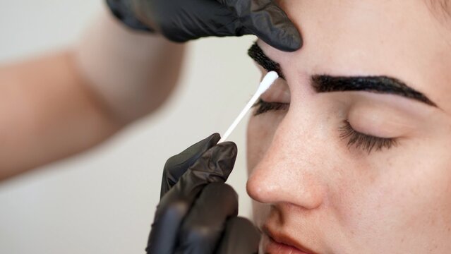 Close-up, The Hands Of The Cosmetologist In Black Rubber Gloves Hold A Cotton Swab And Correct The Shape Of The Eyebrows During Painting. Coloring, Eyebrows Shaping. High Quality Photo