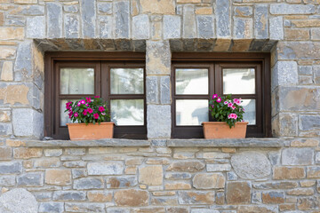 Two symmetric windows with brown pots with pink and purple flowers in a grey stone window from a rural house