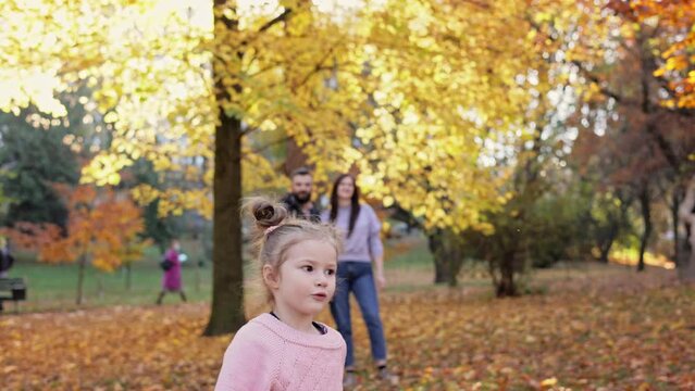 Happy little girl playing with toy airplanes in autumn park, parents looking at her and smiling.Young family enjoy spending time together.Parenthood, carefree childhood, love and happy family concept.