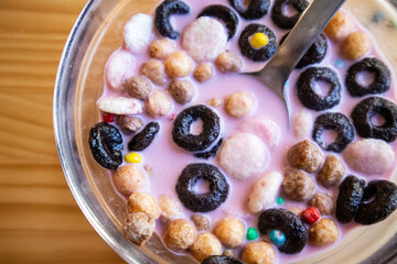 Dry breakfast in glass round plate bowl on a wooden table in cafe. Delicious cornflakes drenched in pink strawberry milk. Healthy breakfast. Food for kids. Different type cereals, multicolored balls.
