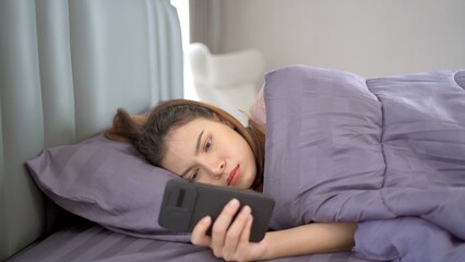 Lazy Asian young woman laying down on the bed and texting in social network with a friends close up.