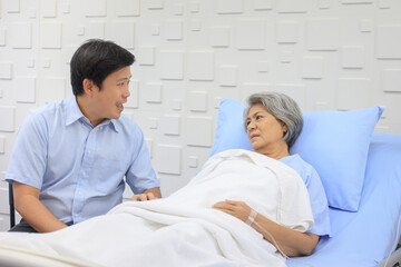 Senior Asian female patient resting on the medical bed in hospital and talking to doctor.