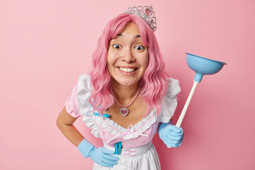 Positive Asian housemaid with pink dyed hair wears crown dress and rubber gloves focused directly...