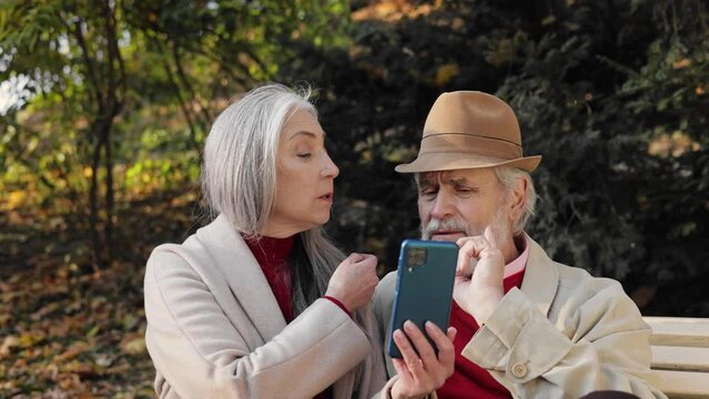 Happy old mature retired couple using smartphone for watching funny photos and video, mobile applications, social media, shopping online. Middle aged spouses enjoying time together in autumn park.