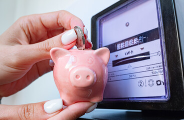 Woman puts coin in piggy bank near a electricity meter. Electricity consumption, cost of utilities...