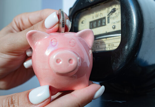 Coin and piggy bank in female's hands near home electricity meter. Symbolic image of energy costs and electricity prices, saving the household budget on paying utility bills.