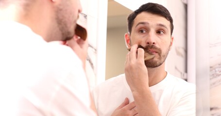 Hipster man focusing on combing beard in front of a mirror.
