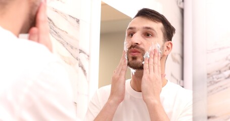 Happy guy is ready to shave. Man Applying Shaving Cream On His Face Standing In Bathroom.