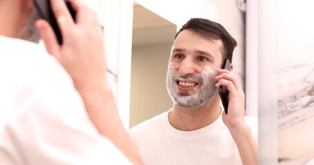 Man talking on a mobile phone in the bathroom. Shave foam on face.