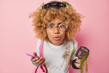 Photo of displeased curly haired woman has dangerous ride on bicycle holds mobile phone and pedal focused at camera with amazed expression wears protective helmet and gloves isolated on pink wall