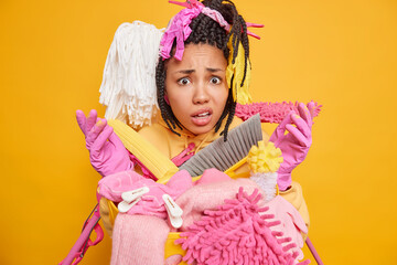 Displeased shocked woman with braided hairstyle has embarrassed expression keeps palms raised wears rubber gloves surrounded by cleaning equipment and detergents isolated over yellow background