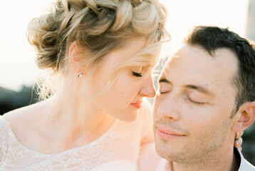 Bride touches groom cheek with her nose, sitting in his arms. Portrait