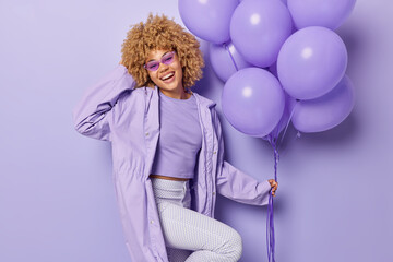 Obraz na płótnie Canvas Cheerful curly haired woman poses with bunch of inflated balloons wears stylish clothes of one color celebrates special occasion comes on party isolated over purple background. Holiday concept